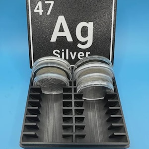 May include: A black tray with compartments holding two stacks of silver coins. Behind the tray is a black sign with the number 47, the letters Ag, and the word Silver in white.