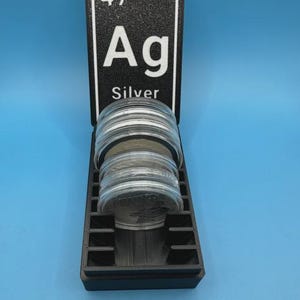 May include: A black display case with a silver periodic table element sign that reads "47 Ag Silver". The case holds several silver coins. The background is a solid light blue.