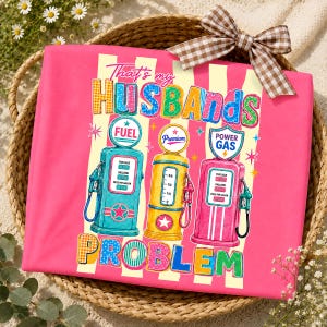 May include: A pink t-shirt with the text "That's my Husbands Problem" and illustrations of vintage gas pumps in teal, yellow, and pink. The shirt has a striped background and is decorated with a brown and white checkered bow.
