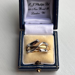 May include: A gold and silver ring in a blue jewelry box with a yellow velvet interior. The ring has a unique, intertwined design. The box is labeled "F.J. Phillips Ltd, 139 New Bond St, London, W1."