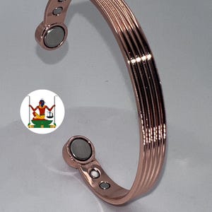 May include: A rose gold-toned metal cuff bracelet featuring a series of parallel vertical ridges along the outer surface. The interior ends of the open-ended bracelet are equipped with two circular magnets and two smaller silver-toned metal studs.