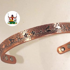 May include: A copper cuff bracelet featuring an embossed desert landscape with cacti and a howling wolf under a moon. The interior of the bracelet displays six small circular magnets embedded at the ends of the band. A small circular logo is visible.