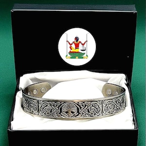 May include: A silver-toned metal cuff bracelet featuring an intricate embossed Celtic knotwork pattern and a central tree motif. The bracelet has visible circular magnets on the interior and is presented in a black box with a white interior lining.