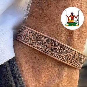 May include: A copper-toned metal cuff bracelet featuring an embossed design of a tree with sprawling branches and intricate interwoven knotwork patterns along the band. The surface has a dark, oxidised finish within the recessed areas of the metal.