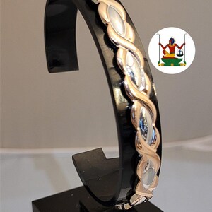 May include: A two-tone bracelet featuring a braided design with alternating rose gold and silver-coloured metallic links. The bracelet is displayed on a black curved stand and includes a circular logo depicting a figure holding a scale.