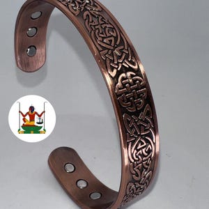 May include: A copper-coloured cuff bracelet featuring an embossed Celtic knot pattern across the outer surface. The interior of the bracelet displays six small, circular silver-toned magnets embedded into the metal at both ends of the open band.