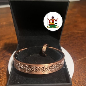 May include: A copper-toned metal cuff bracelet and a matching ring featuring an intricate embossed Celtic knot pattern. The jewellery is displayed inside a black velvet-lined box, which also contains a circular sticker depicting a seated figure.
