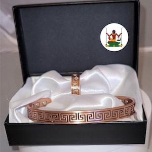 May include: A copper-coloured metal cuff bracelet and a matching ring, both featuring a repeating black Greek key pattern. The bracelet has visible circular magnets on the interior. The items are presented inside a black box with white fabric.