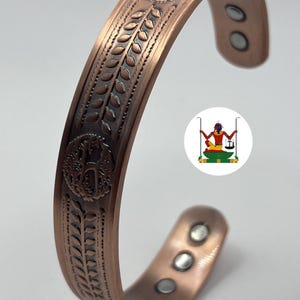 May include: A copper-coloured cuff bracelet featuring embossed tree and leaf motifs along the outer surface. The interior of the open-ended band displays six circular silver-toned magnets embedded at the ends. A small circular logo is visible.