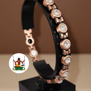 May include: A bracelet featuring a black band accented with copper-toned metal links. Each link is set with a clear, round, faceted stone. The interior of the band displays small black and white circular elements. A circular logo is visible on the left.