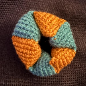 May include: A handmade crochet donut-shaped item. The item is composed of alternating sections of teal and orange yarn. The yarn is tightly crocheted, creating a textured surface. The item is displayed against a dark, neutral background.