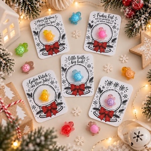 May include: Five white rectangular cards featuring a black line drawing of a snow globe with a red bow, each holding a small translucent turtle figurine. The cards display text including Merry Christmas, Frohes Fest, Guten Rutsch, and HO-HO-HO.