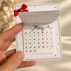 May include: A white paper calendar for the month of M&auml;rz featuring a grid of dates from 1 to 31. The top left corner is decorated with a small red ribbon bow and a ladybug charm. The calendar has a black border with small dots.