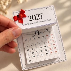 May include: A 2027 wall calendar with a white background, black text, and a red ribbon bow featuring a small ladybug embellishment. The top page displays the text '2027' and 'Jeder Tag ist ein besonderer Tag!', while the page below shows the month of May.