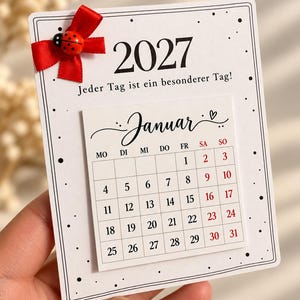 May include: A white 2027 calendar card featuring the text 'Jeder Tag ist ein besonderer Tag!', a red ribbon bow, and a small ladybug embellishment. The calendar displays the month of 'Januar' with a grid of dates in black and red ink.
