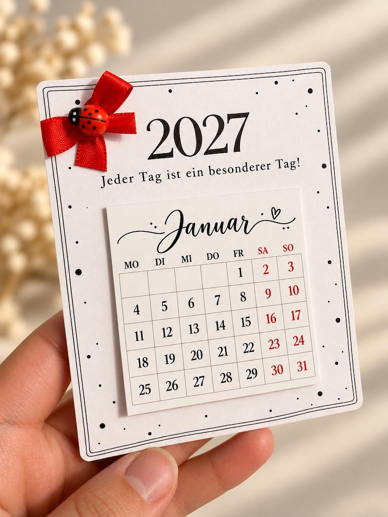 May include: A white 2027 calendar card featuring the text 'Jeder Tag ist ein besonderer Tag!', a red ribbon bow, and a small ladybug embellishment. The calendar displays the month of 'Januar' with a grid of dates in black and red ink.