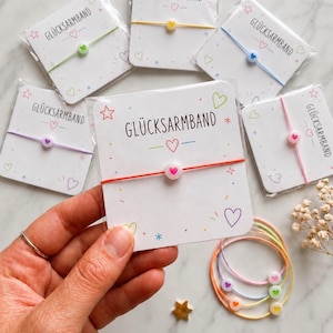 May include: A collection of thin string bracelets, each featuring a single round bead with a small heart print. The bracelets are individually packaged on white cards labeled GLÜCKSARMBAND with colorful star and heart illustrations.