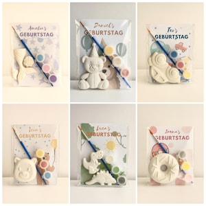 Op de afbeelding: Doe-het-zelf knutselsets om te schilderen, met vormen zoals een zeemeerminstaart, teddybeer, auto, dinosaurus en donut. Elke set bevat een penseel en kleine verfpotjes. De verpakking bevat de tekst "Amelia's GEBURTSTAG", "Daniel's GEBURTSTAG", "Teo's GEBURTSTAG", "Icon's GEBURTSTAG", "Luca's GEBURTSTAG" en "Irena's GEBURTSTAG".