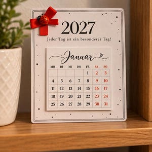 May include: A rectangular 2027 calendar card featuring the text 'Jeder Tag ist ein besonderer Tag!' and a January grid. The card is decorated with a red ribbon bow and a small ladybug embellishment, set against a white background with black dots.