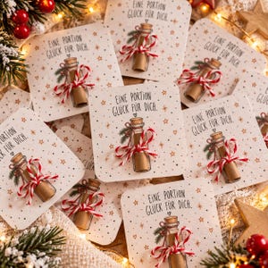 May include: A collection of square white cards featuring a printed illustration of a glass bottle with a cork, tied with a red and white string, and the German text "Eine Portion Glück für dich" surrounded by small stars and hearts.
