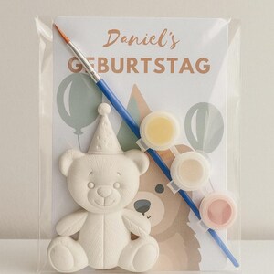 Könnte beinhalten: Ein Mal-dein-eigenes-Bären-Set in einer transparenten Kunststoffverpackung. Das Set enthält einen weißen Keramikbären mit Partyhut, drei Farbtöpfe und einen Pinsel. Die Karte trägt den Text "Daniel's GEBURTSTAG" und eine Bärenillustration.