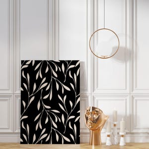 May include: A black and white canvas print featuring a repeating pattern of stylized leaves. The artwork is displayed against a white paneled wall, with a gold-colored sculpture of a face, candles, and a hanging light fixture.