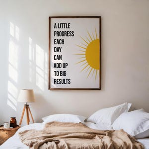 Puede incluir: Un estampado enmarcado con el texto "A LITTLE PROGRESS EACH DAY CAN ADD UP TO BIG RESULTS" y un gráfico de sol amarillo. El estampado está en un marco marrón y cuelga sobre una cama con almohadas blancas y una manta beige. Una lámpara con base de madera está cerca.