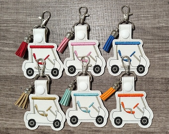 May include: Six white golf cart keychains with colorful tassels. Each keychain features a white golf cart with a red, blue, orange, pink, light blue, or green tassel.
