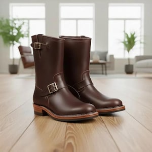 May include: A pair of brown leather boots with a mid-calf height. The boots feature a buckle closure and a stacked heel. The soles have a light brown border. The boots are displayed on a wooden floor.