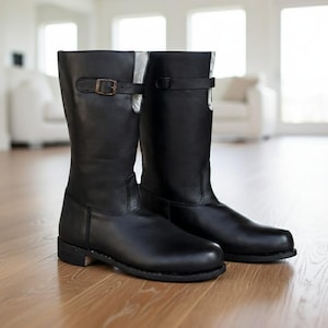 Könnte beinhalten: Ein Paar schwarze Lederstiefel, Wadenmitte, mit Schnallenverschlüssen. Die Stiefel werden auf einem Holzboden präsentiert. Das Design ist klassisch und praktisch, geeignet für verschiedene Wetterbedingungen. Die Höhe beträgt ca. 30 cm.