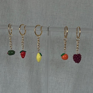 Puede incluir: Cinco llaveros con anillos y cadenas doradas, cada uno con un dije de fruta diferente: una aceituna, una fresa, un limón, una naranja y una mora. Los dijes son de colores brillantes y detallados.