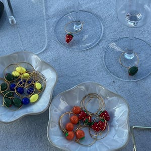 Puede incluir: Un conjunto de adornos para copas de vino y cuencos decorativos. Los adornos presentan frutas en miniatura como fresas, naranjas y aceitunas, unidas a aros dorados. Los cuencos blancos tienen un borde festoneado, sobre un mantel azul claro.