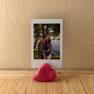 May include: A glossy, pink heart-shaped ornament sits on a wooden surface. Behind it is a Polaroid photo of a couple kissing, holding a sonogram. The background shows a lake and trees.