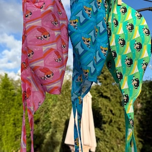 May include: Three colorful head wraps hanging on a clothesline. The wraps feature cartoon character prints in pink, blue, and green. The pink wrap has a repeating pattern of a character with orange and purple accents. The blue wrap has a repeating pattern of a character with blue and yellow accents. The green wrap has a repeating pattern of a character with black and yellow accents.