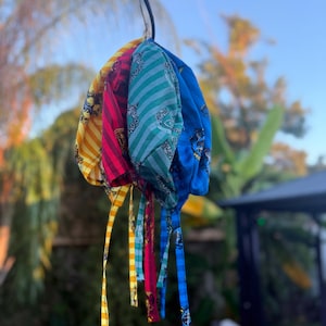 Könnte beinhalten: Eine Sammlung farbenfroher Kopftücher, jedes mit einem einzigartigen Muster. Die Tücher hängen an einem Ast und zeigen eine Vielzahl von Designs, darunter Streifen und Paisley, in Gelb-, Rot-, Türkis- und Blautönen. Die Bänder sind ebenfalls gemustert.