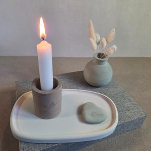 Könnte beinhalten: Eine brennende weiße Kerze in einem Betonhalter steht auf einem weißen Tablett mit einem herzförmigen Stein. Eine kleine, runde, graue Vase enthält Trockenblumen. Die Artikel liegen auf einer grau strukturierten Oberfläche und schaffen eine minimalistische Ästhetik.