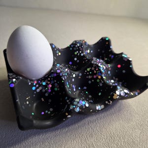 Könnte beinhalten: Ein schwarzer Eierhalter mit einem gesprenkelten, glitzernden Design, der ein einzelnes weißes Ei hält. Der Eierhalter hat eine wellenförmige, geformte Form. Der Glitzer enthält schillernde Farben.