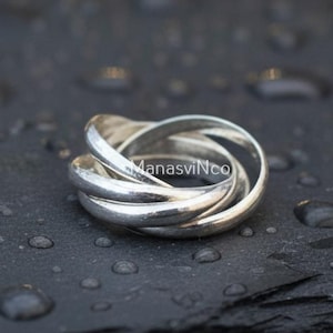 Könnte beinhalten: Ein silberner Ring mit einem dreibändigen Design, der auf einer dunklen, strukturierten Oberfläche liegt. Die Bänder des Rings sind ineinander verschlungen, was einen einzigartigen Look erzeugt. Ein Schmuckstück.