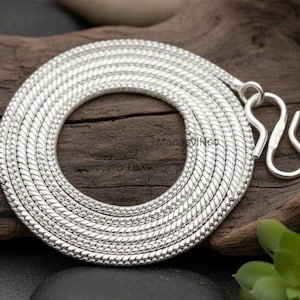Può includere: Una collana argentata arrotolata con un design strutturato a forma di serpente. La collana è appoggiata su una superficie di legno e presenta una chiusura a gancio. Il nome del marchio "ManasviNco" è visibile sul legno.