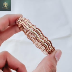 May include: A stack of copper-colored bangles with a wavy design. The bangles are held in a hand, showcasing their intricate, layered construction. The metal has a warm, reddish-brown hue, with hints of gold.