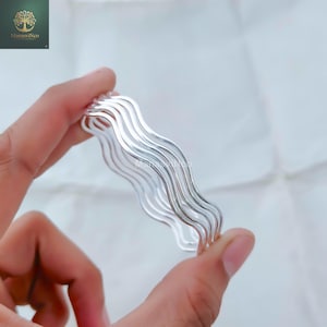Può includere: Un gruppo di braccialetti argentati con bordi ondulati. I braccialetti sono tenuti in una mano, evidenziando la loro superficie liscia e riflettente. Il design è semplice ed elegante, adatto per l'uso quotidiano o occasioni speciali.
