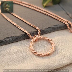 May include: Rose gold necklace featuring a braided circle pendant. The chain and pendant have a matching metallic finish. The necklace is displayed on a neutral-colored surface, showcasing the jewelry's design and color.