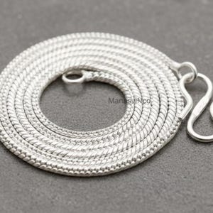 May include: A coiled silver snake chain necklace with a hook clasp. The chain is made of small, tightly linked segments, creating a textured, flexible design. The necklace is displayed on a grey surface.