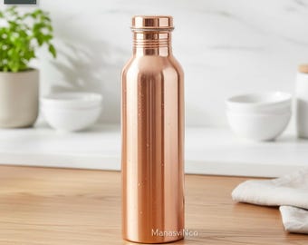 Puede incluir: Una botella de agua de color cobre con tapón de rosca. La botella es cilíndrica y tiene una superficie lisa y reflectante. La botella está sobre una superficie de madera.