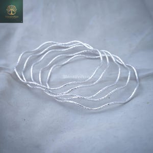 May include: A collection of silver bangles with a wavy, textured design. The bangles have a hammered finish and a fluid, organic shape. The background is a soft, neutral colour. The brand name "ManasvinCo" is visible in the image.