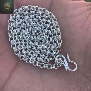 Puede incluir: Un collar de cadena plateada con cierre de gancho. La cadena está hecha de eslabones ovalados entrelazados. El collar se muestra en una mano. La joyería es probablemente para adorno personal.