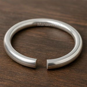 Puede incluir: Un brazalete abierto de color plateado. El brazalete tiene un diseño liso y redondeado y está hecho de un metal brillante. El texto "ManasVinco" es visible en el brazalete. El brazalete se muestra sobre una superficie de madera marrón oscuro.