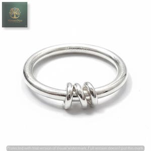 Puede incluir: Un anillo de plata con una banda gruesa y tres anillos más pequeños conectados. El anillo se muestra sobre un fondo blanco. La marca "ManasviNco" es visible en la esquina superior izquierda.