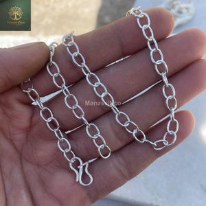 May include: A silver-colored chain necklace with oval links and a hook clasp. The necklace is displayed on a hand. The brand name "ManasviNco" is visible in the upper left corner.