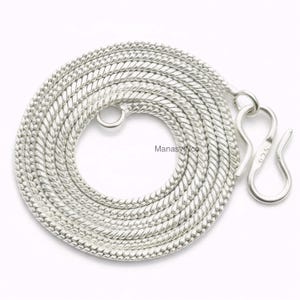 以下が含まれることがあります： 織り込まれたテクスチャデザインの、銀色のコイル状ネックレス。 ネックレスはフッククラスプと端に小さなリングが付いています。 ジュエリーは白い背景に表示されています。